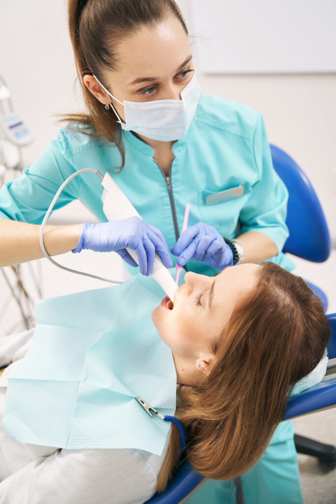 Dentist in medical mask performing intraoral scanning procedure with modern equipment while woman lying in dental chair